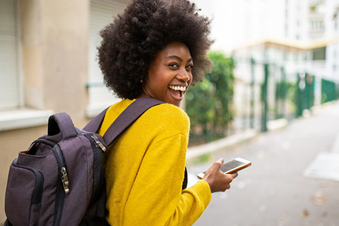 A young woman wearing a yellow sweater and purple backpack holding her phone and smiling in Orange